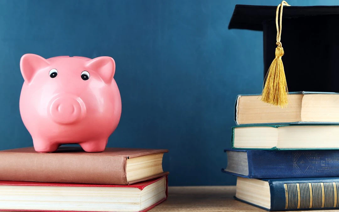 A photo of a piggy bank atop a stack of books with another stack of books and a graduation cap next to it