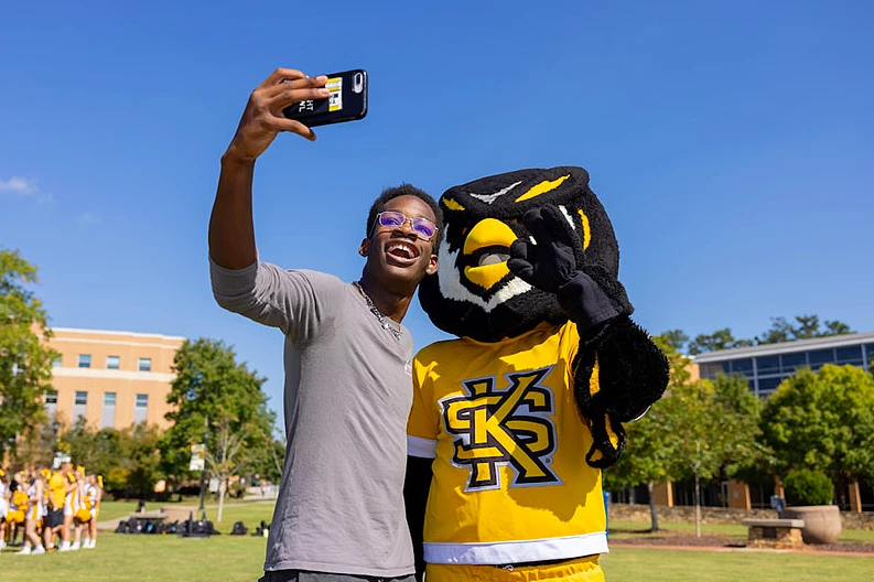 A man holds his phone up to take a picture with an owl mascot