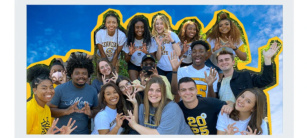 A group of smiling young people pose together outdoors, some wearing shirts with "Kennesaw State" on them.