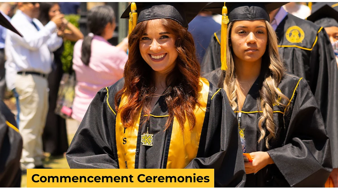 Two graduates in caps and gowns smile and stand together at a commencement ceremony.