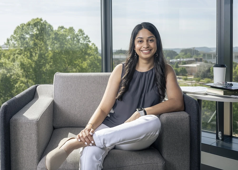 Maya Patel is sitting on a gray armchair by a large window, smiling, with books and a coffee cup on a table beside her.