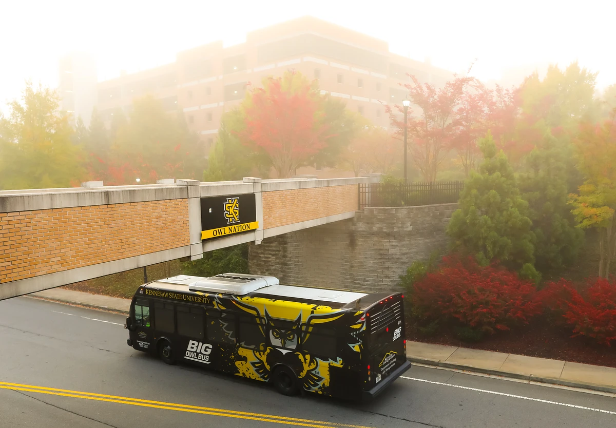 A bus with vibrant university branding drives under a pedestrian bridge labeled "OWL NATION" amidst a foggy backdrop with autumnal trees.